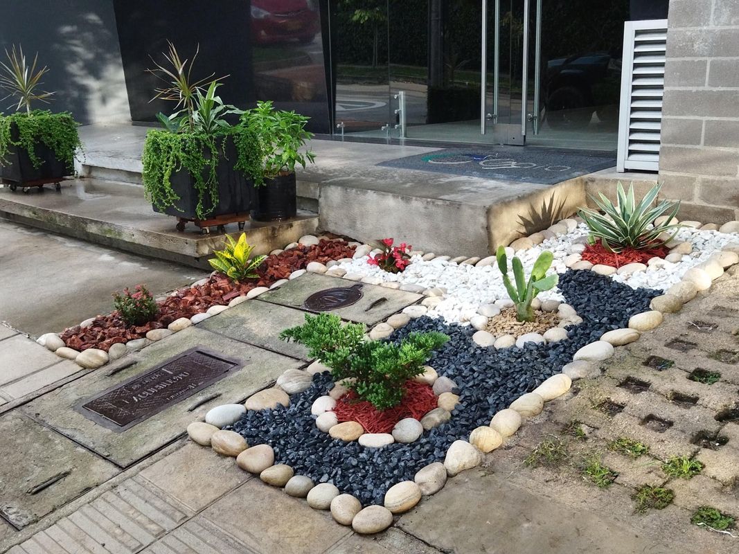 Jardín en la entrada de un edificio con piedras de colores, agave, cactus y materas con plantas colgantes