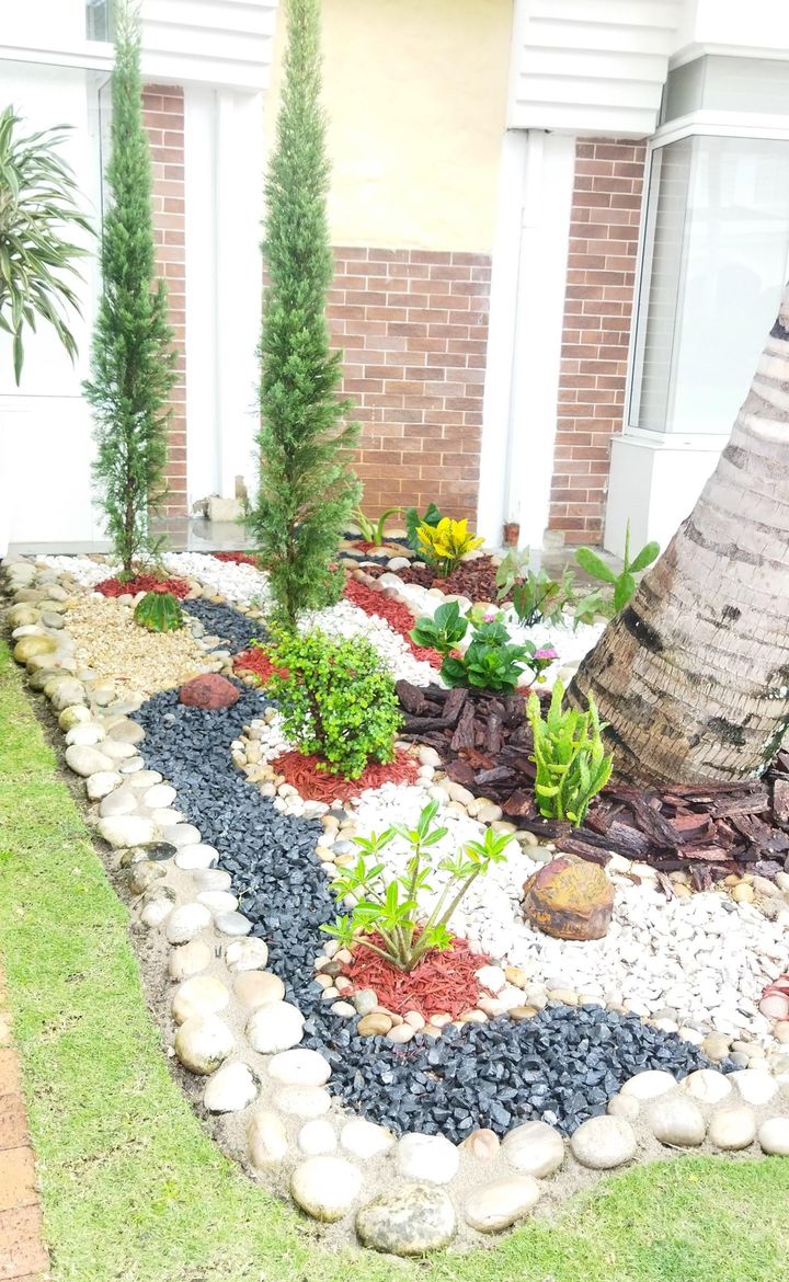 Jardín con cipreses, croto, hortensia y caminos de piedra negra, blanca y corteza roja junto a una palma