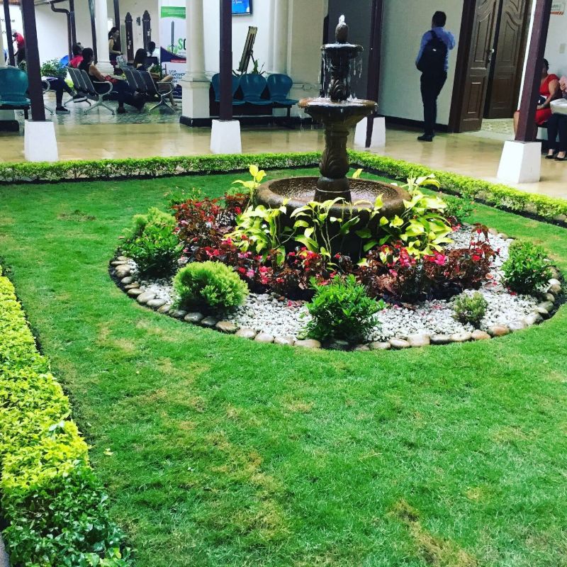 Jardín con césped, setos y una fuente central rodeada de flores y piedra blanca