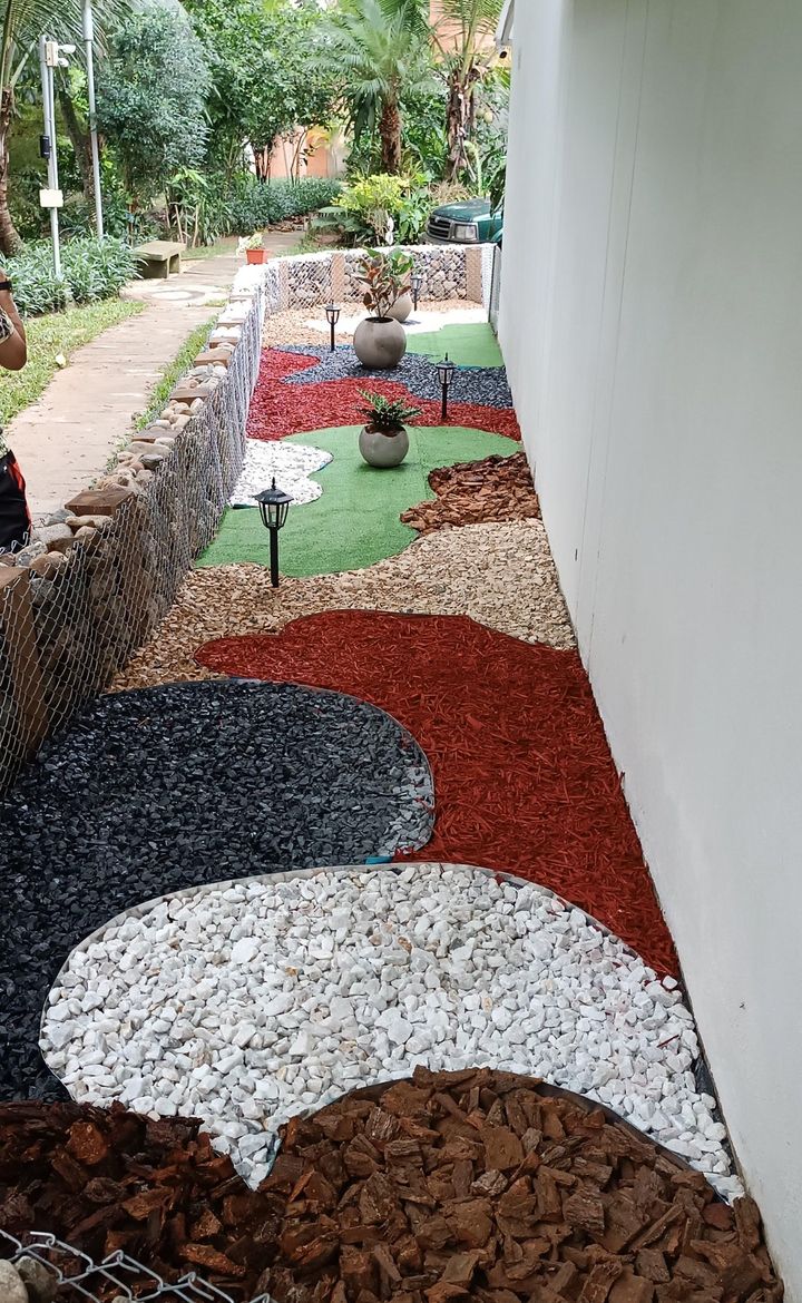 Pasillo lateral con gravas de colores, corteza roja, prado sintético, materas esféricas y faroles