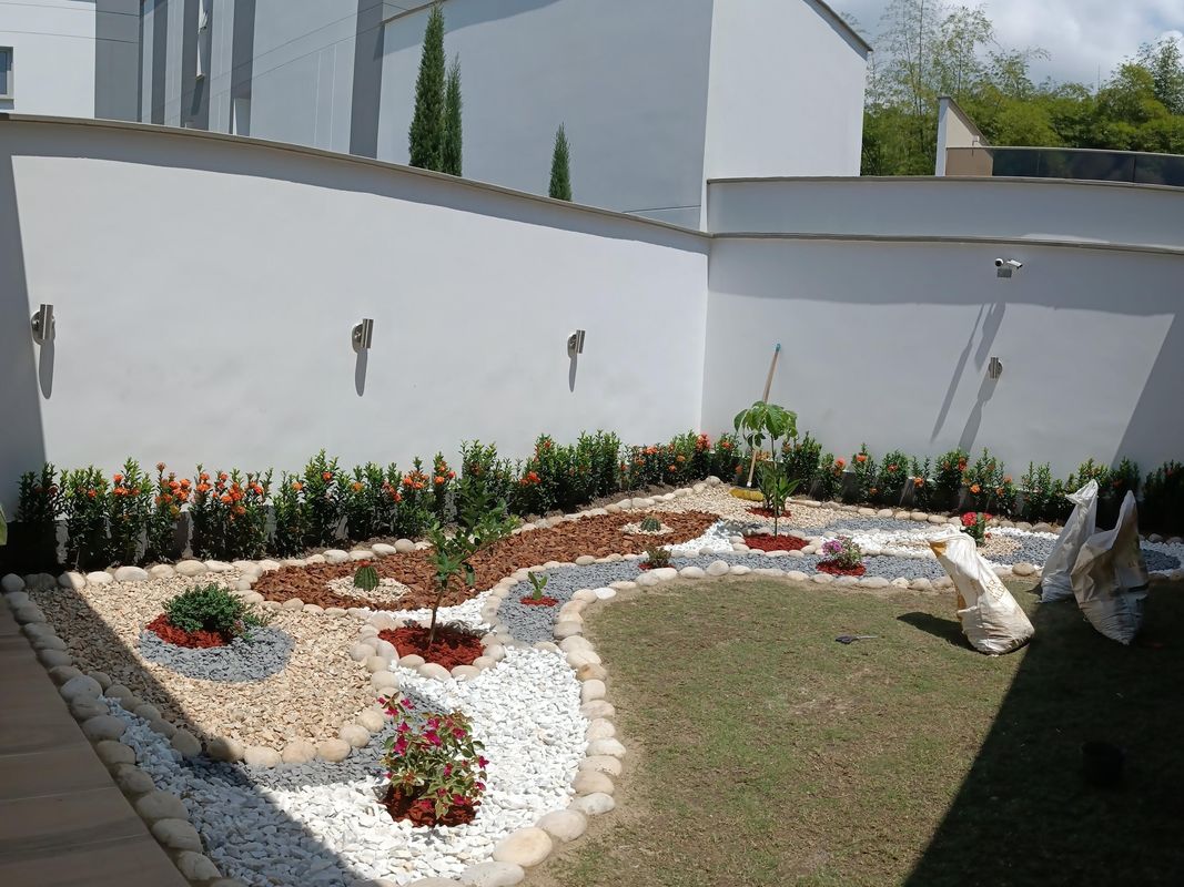 Patio con diseño de piedras blancas, grises y corteza, bordes de piedra de río y una hilera de ixoras naranjas junto al muro